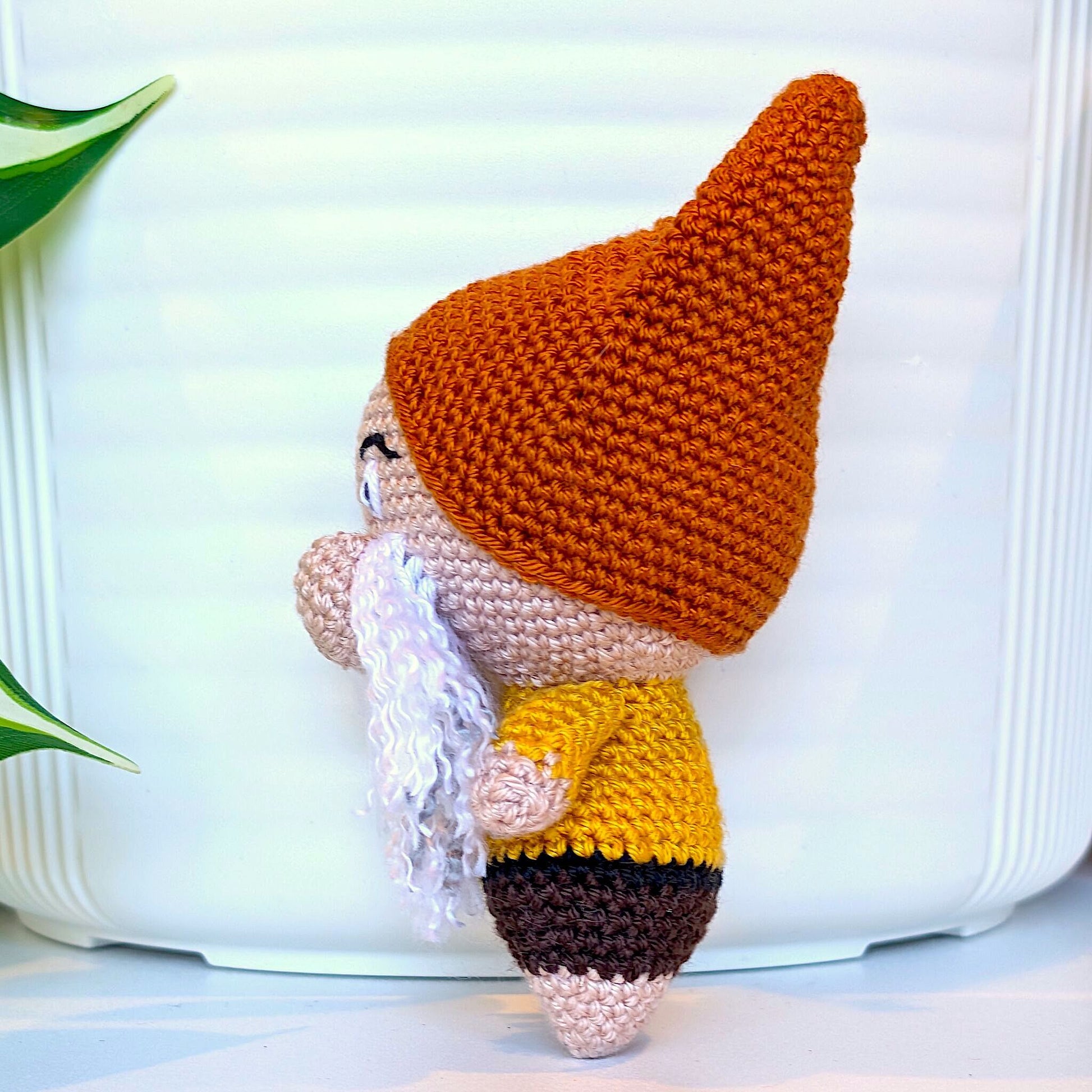 A handmade crocheted gnome doll with a pointed terracotta brown hat, golden yellow shirt, and white beard, standing
