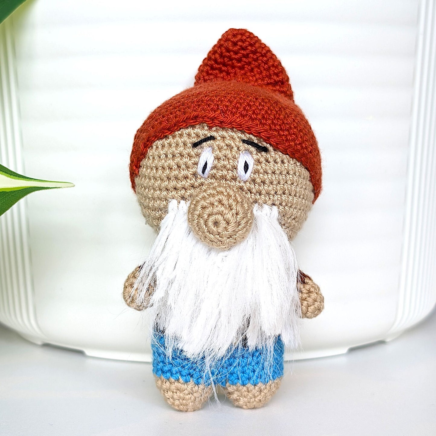 Handmade crocheted gnome figurine toy with happy expression, white beard, mahogany colored hat and brilliant azure blue pants