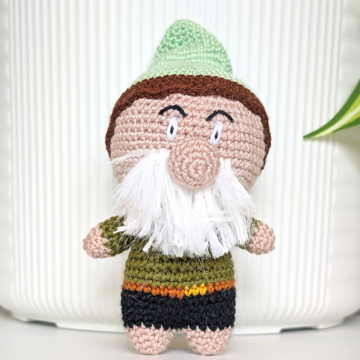 A handmade crocheted gnome doll with a mint green hat with brown trim, a white beard, and a green, and black outfit, standing in front of a white ribbed background.