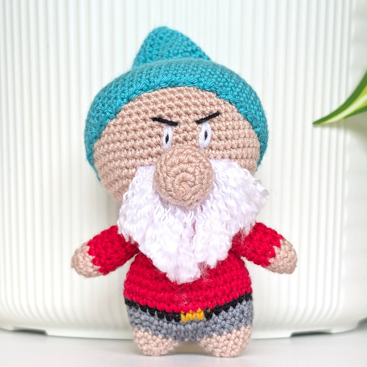 A handmade crocheted amigurumi doll of a gnome character, featuring a teal hat, beige face and arms, a white beard, a rose shirt, and gray pants, set against a white ribbed background.