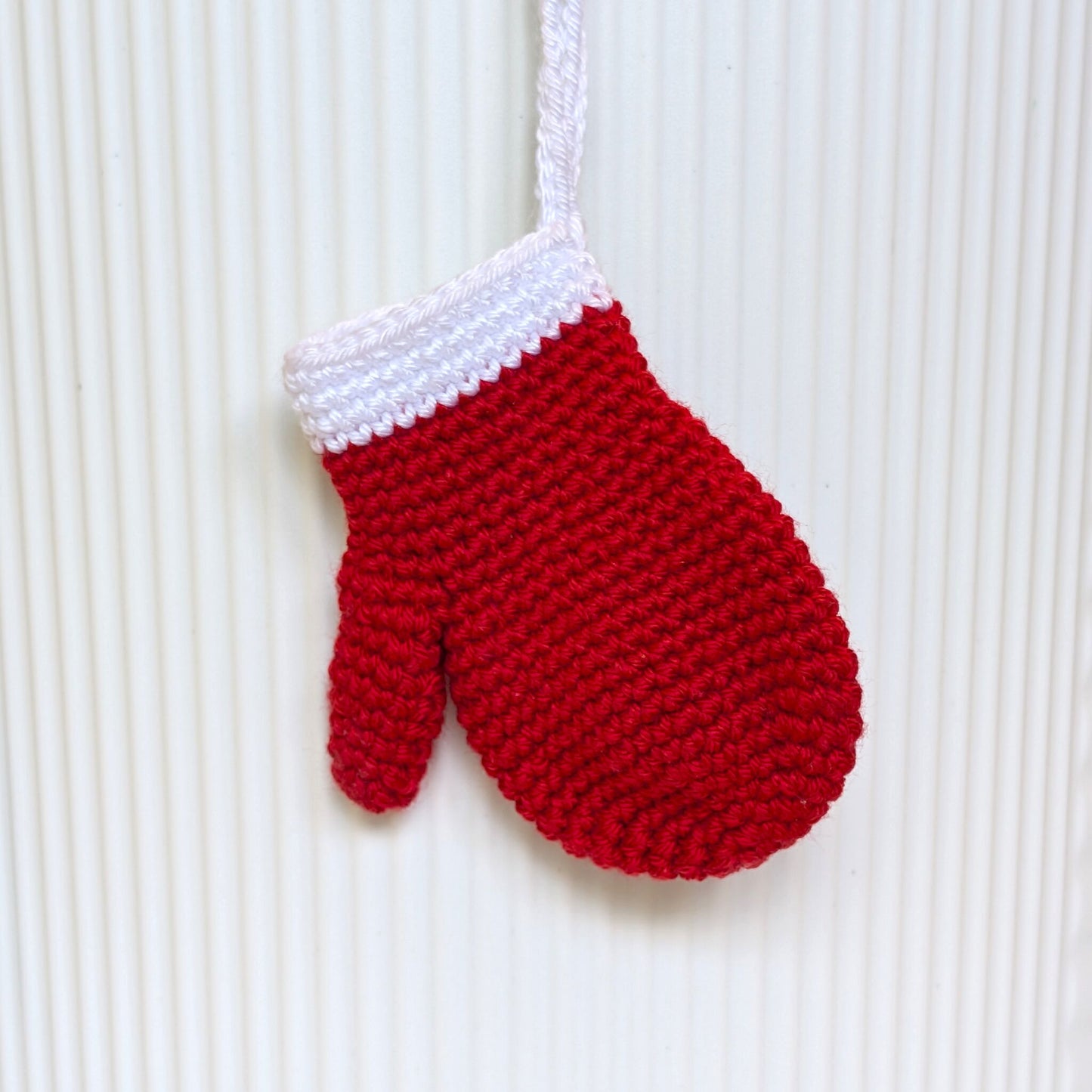 A red and white crocheted mitten ornament hanging against a white ribbed background.