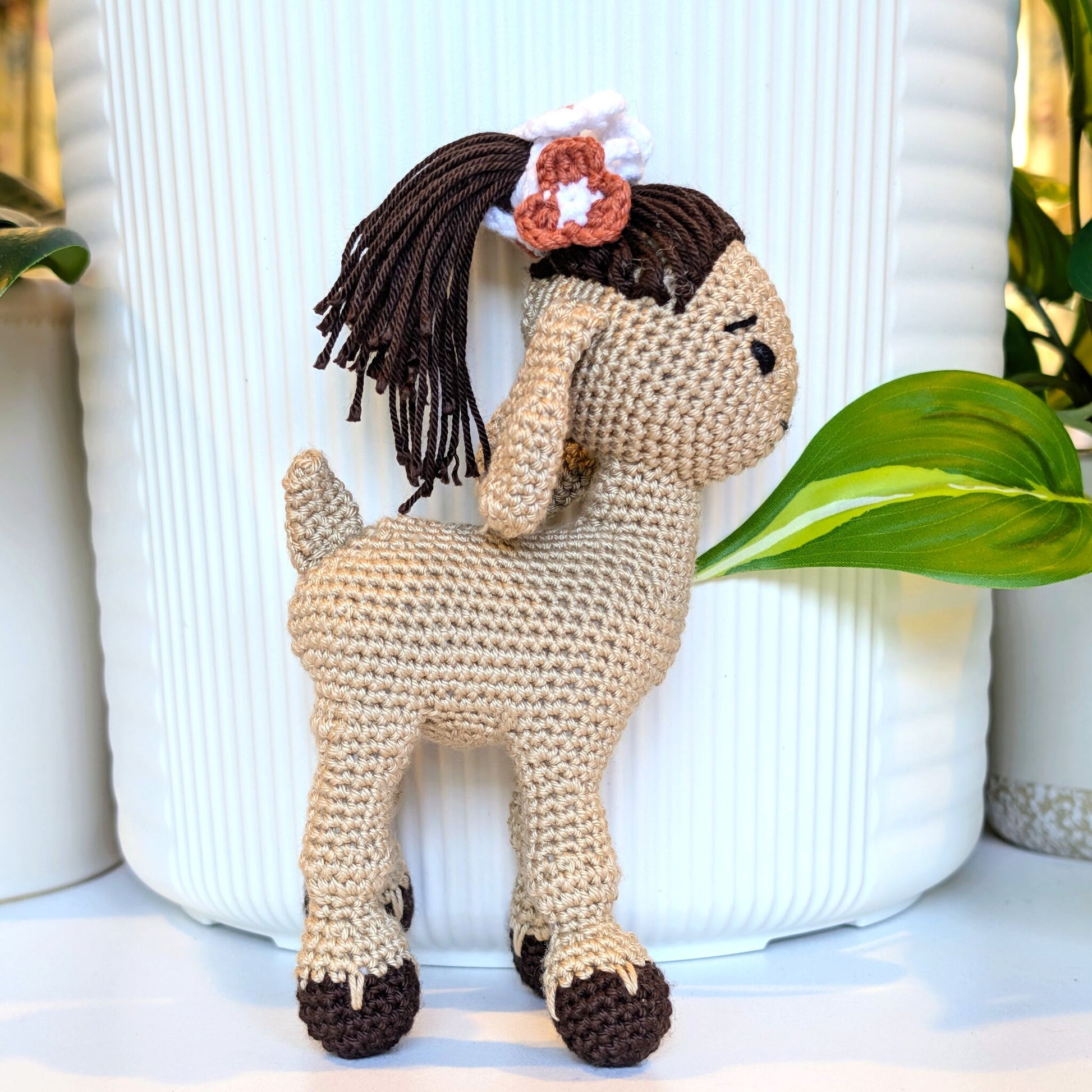 A handmade crocheted stuffed goat with dark brown yarn hair and dark brown hooves, posed in front of a white ribbed ceramic pot with green leaves in the background.