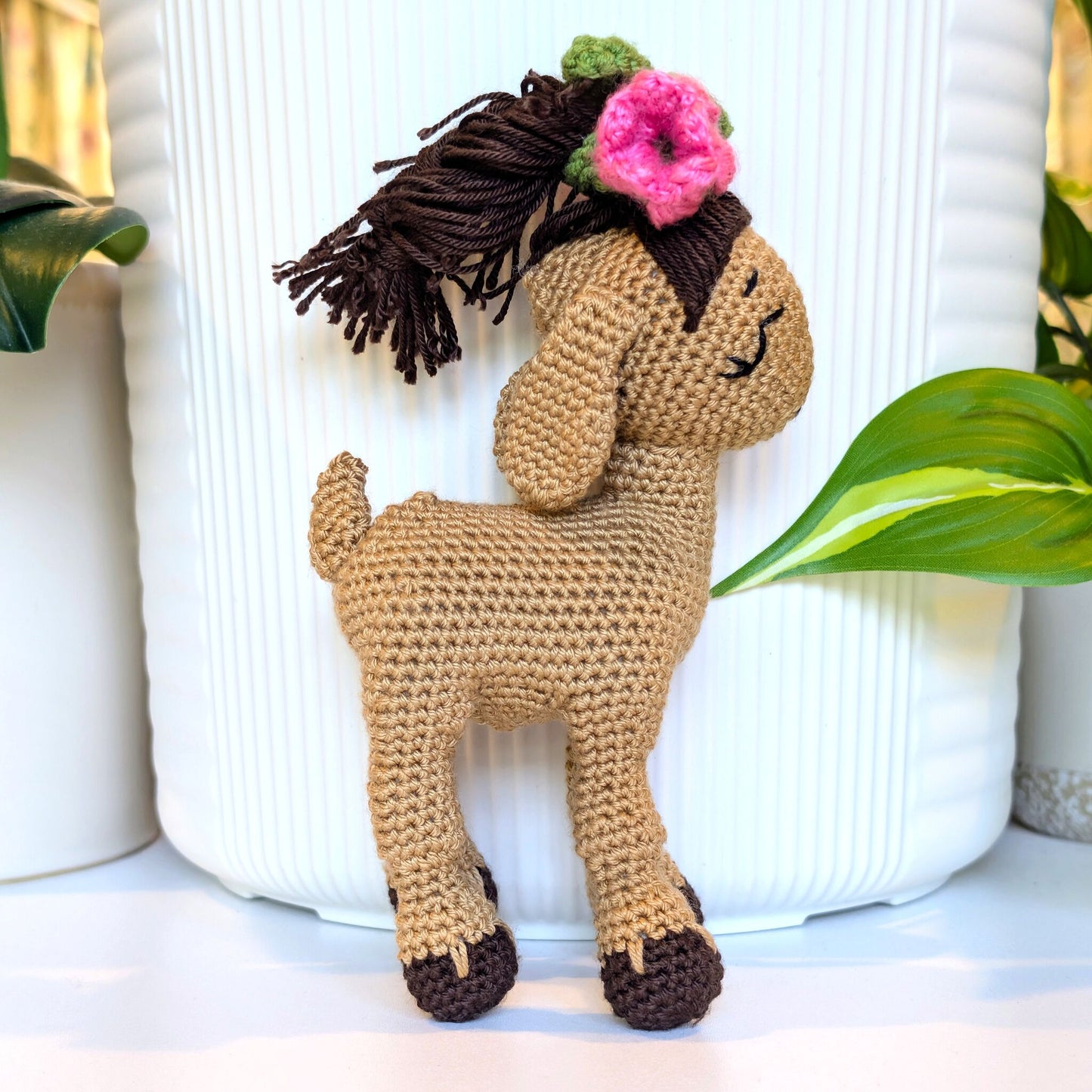 A crocheted stuffed goat, standing in front of a white ribbed vase. The toy has a tan body, dark brown hair with a pink flower and green leaves, and dark brown hooves.
