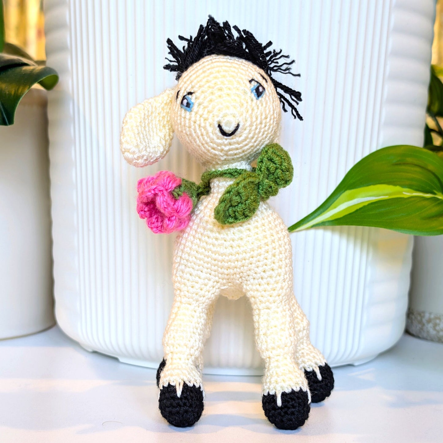 A crocheted stuffed animal goat figure with cream-colored body, black yarn hair, blue embroidered eyes, a green leafy collar, and a bright pink flower in one ear, posed in front of a white ribbed vase with green leaves around it.