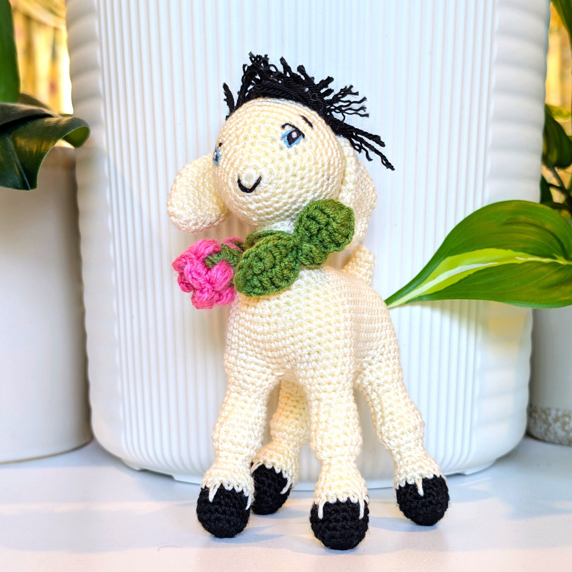 A crocheted stuffed animal goat with black yarn hair, a green scarf, and pink flowers in its mouth, standing in front of a white ribbed vase with green plants in the background.