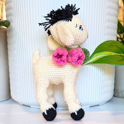 A crocheted plush toy goat with cream-colored body, black yarn hair, and pink flowers on its neck, posed in front of a white ribbed ceramic pot.