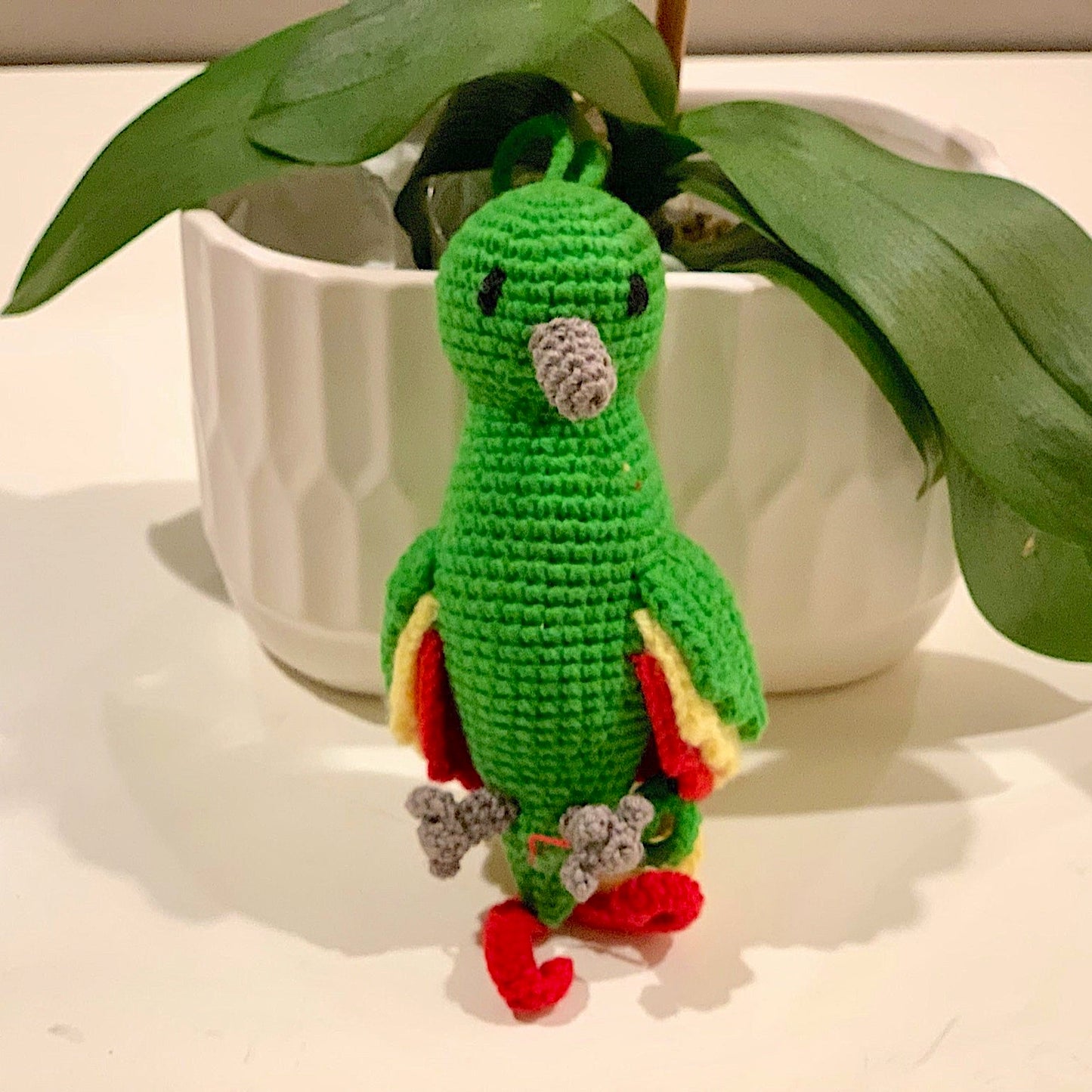 Crocheted green parrot with multi-colored tail feathers and wings on white surface