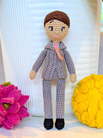 Crocheted boy doll in a grey suit with a pink bow tie and black shoes