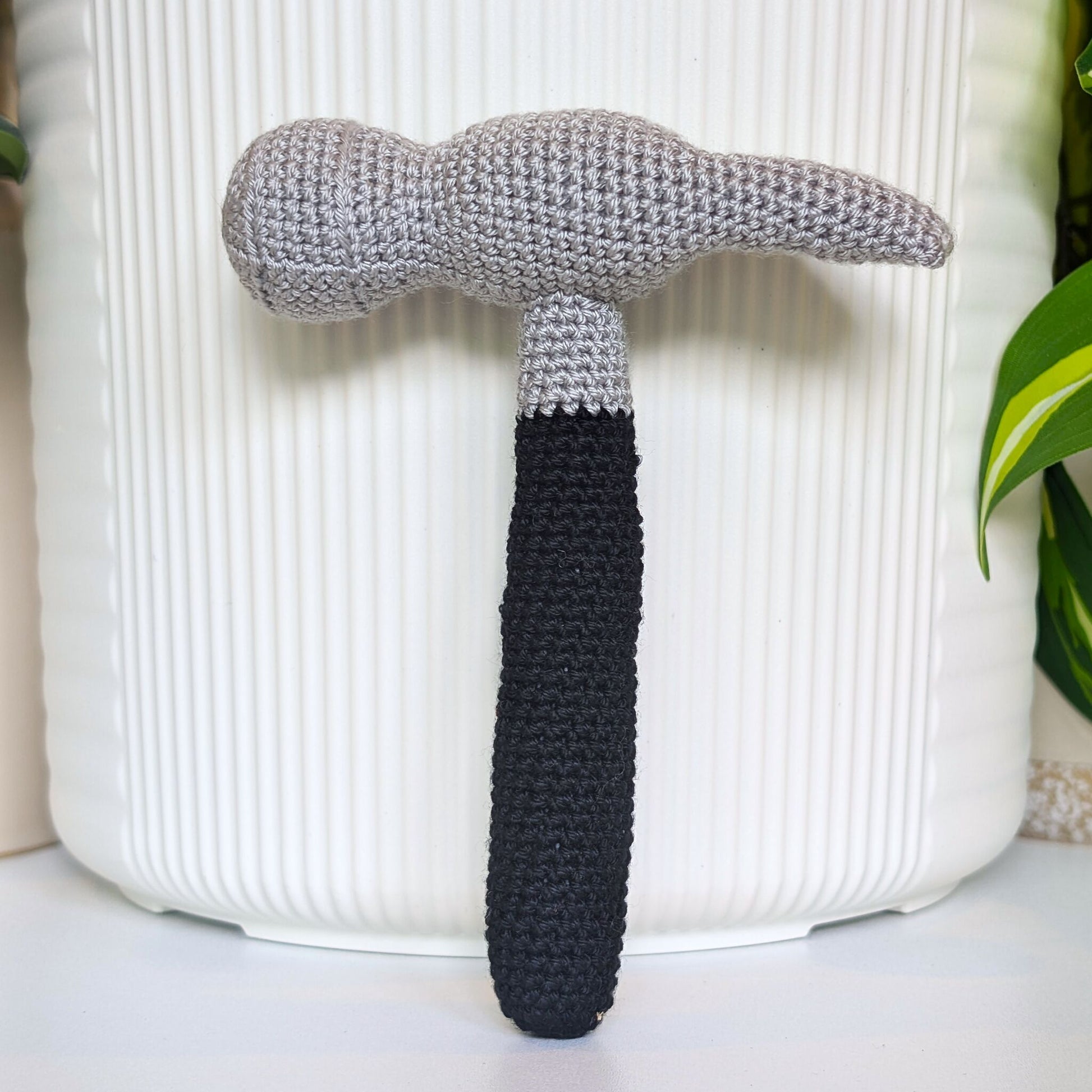 A crocheted hammer with a light gray head and a black handle, standing upright in front of a white ribbed cylindrical planter pot.
