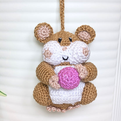 Crochet sand brown hamster bag charm with a string holding an orchid colored ball