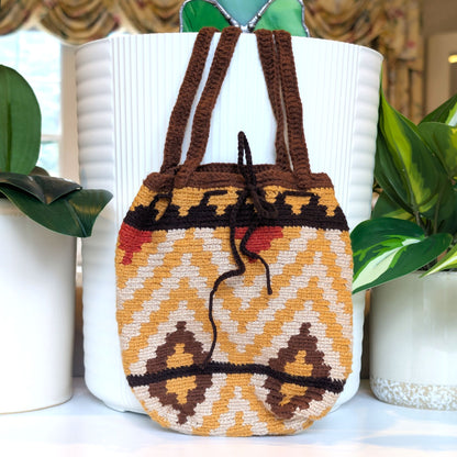 A crocheted hand bag with a Southwestern geometric pattern in mustard yellow, brown, and cream, displayed on a white ribbed planter with green plants in the background.
