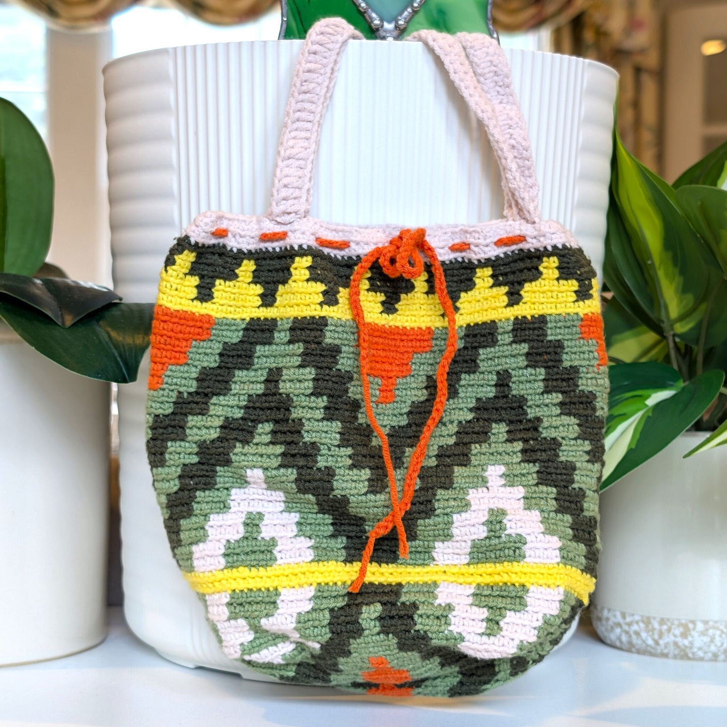 A crocheted handbag with a colorful geometric pattern, featuring green, yellow, orange, and white motifs, displayed in front of a white ribbed planter with green plants.