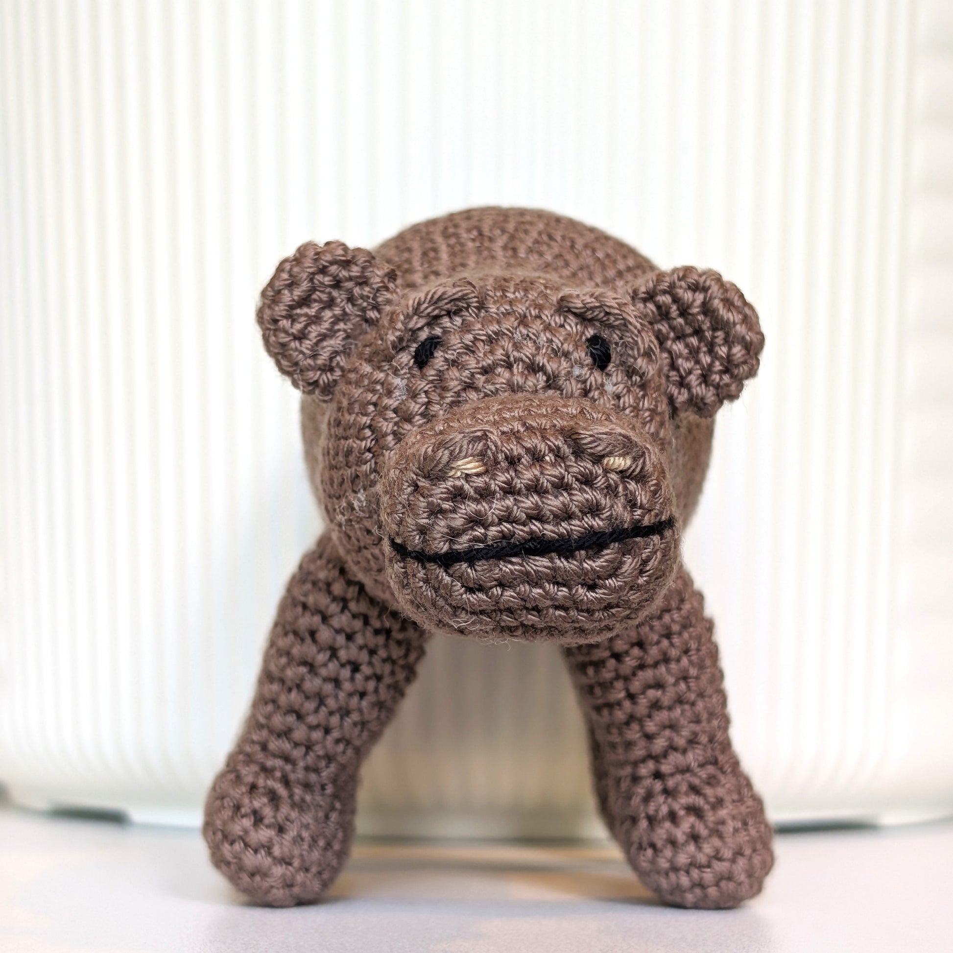A handmade crocheted hippopotamus toy in a medium brown color, standing on a white surface in front of a white ribbed ceramic container.