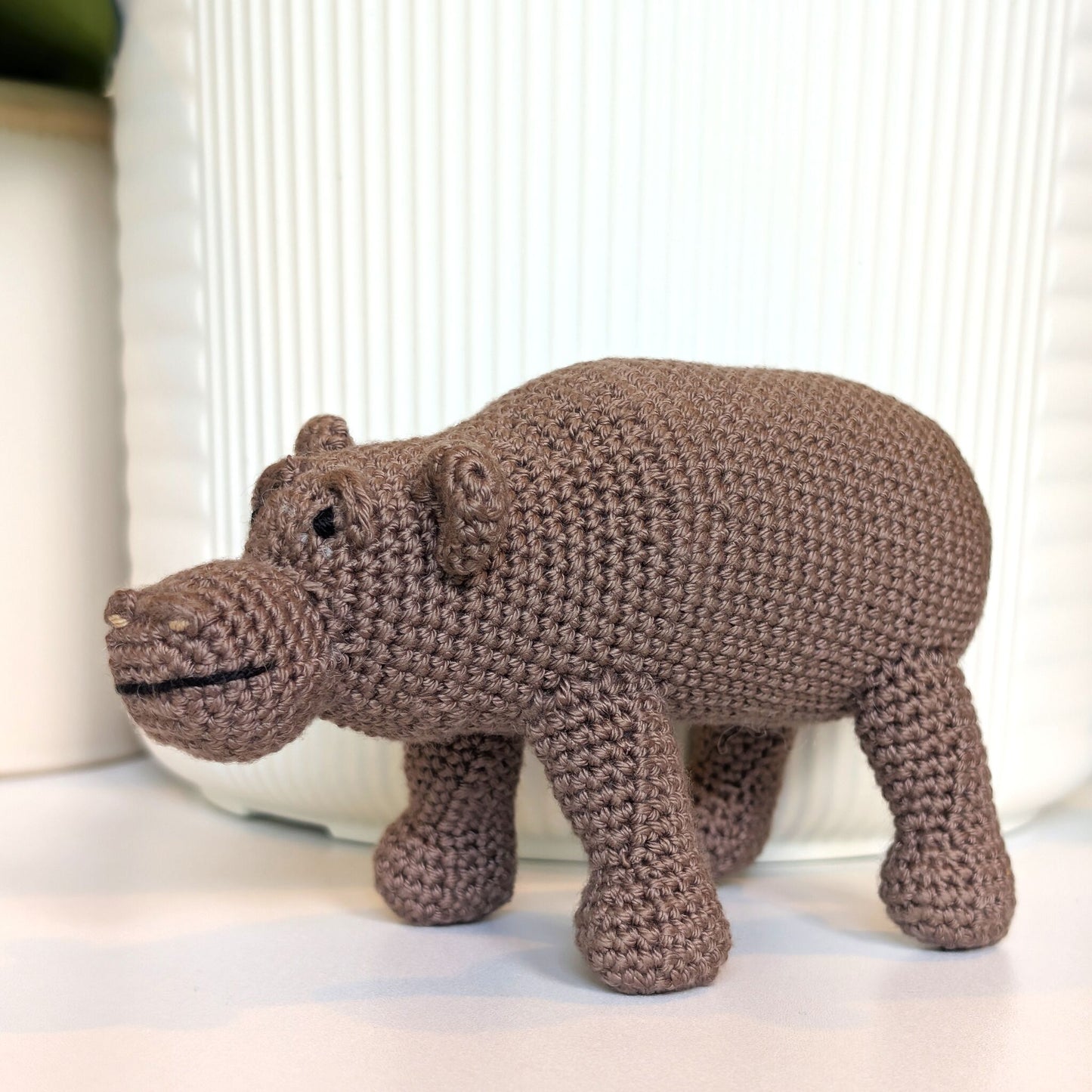 A handmade crocheted hippopotamus toy in a medium brown color, standing on a white surface in front of a white ribbed ceramic container.