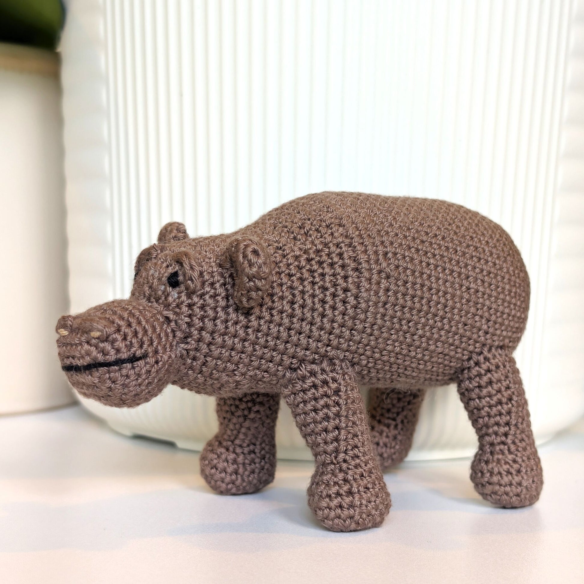A handmade crocheted hippopotamus toy in a medium brown color, standing on a white surface in front of a white ribbed ceramic container.