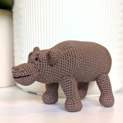 A handmade crocheted hippopotamus toy in a medium brown color, standing on a white surface in front of a white ribbed ceramic container.