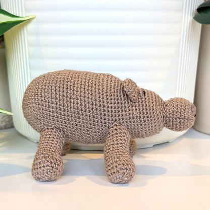 A handmade crocheted hippopotamus toy in a rich taupe color, standing on a white surface in front of a white ribbed ceramic container.