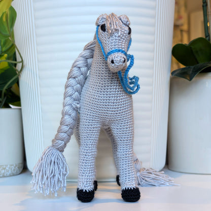 A crocheted plush horse toy with a bright gray body, white mane and tail, black hooves, and a bright blue bridle, standing in front of a white ribbed vase with potted plants in the background.