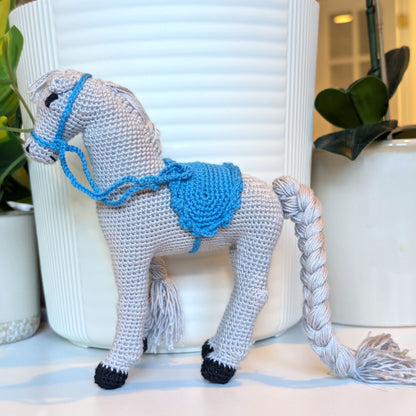 A crocheted toy horse in bright gray yarn with a bright blue saddle and bridle, standing in front of a white ribbed ceramic planter on a tabletop with potted plants in the background.