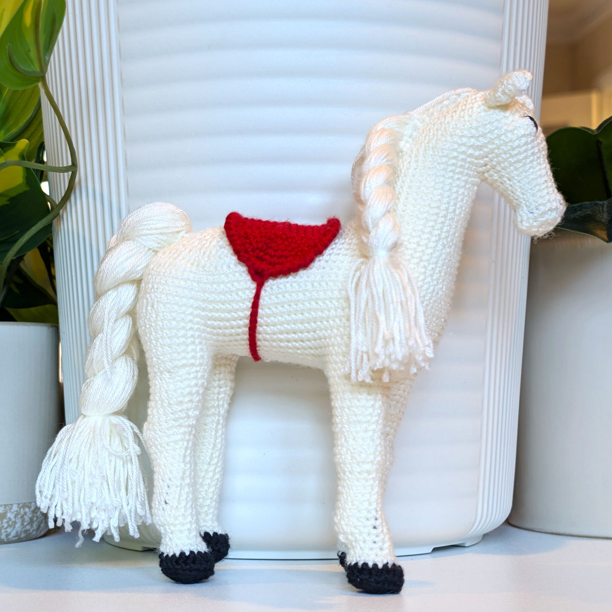 A crocheted horse figurine in off-white with a red saddle and black hooves, standing in front of a white ribbed cylindrical container with potted plants on either side.
