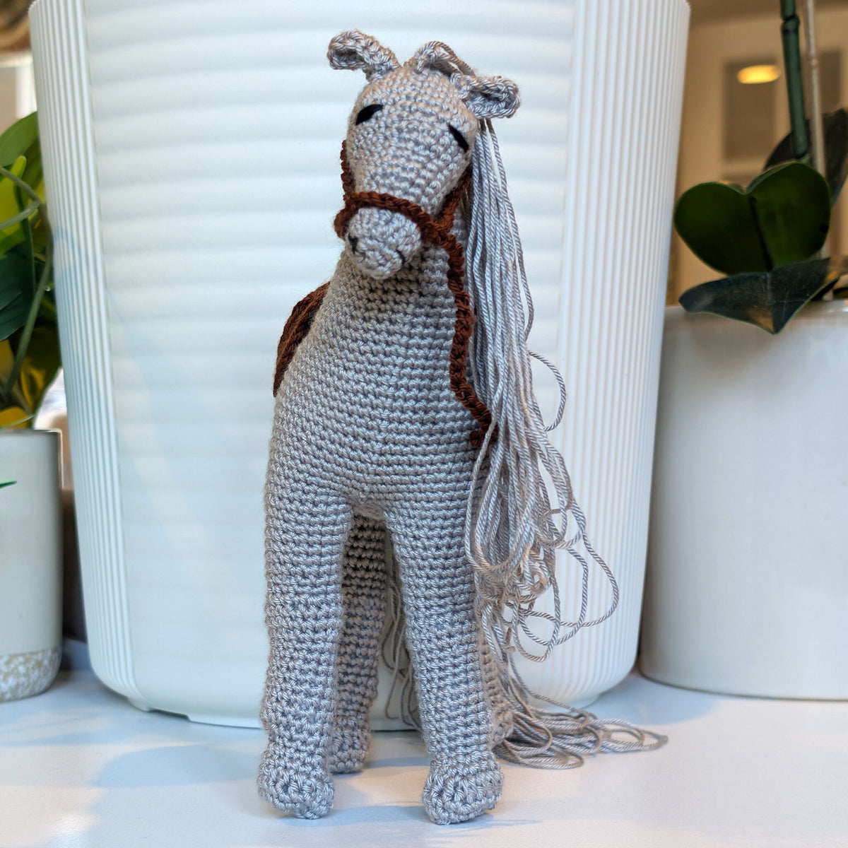 A crocheted stuffed horse toy with a gray body, brown bridle, and long gray mane, standing in front of white ribbed planters with green plants.
