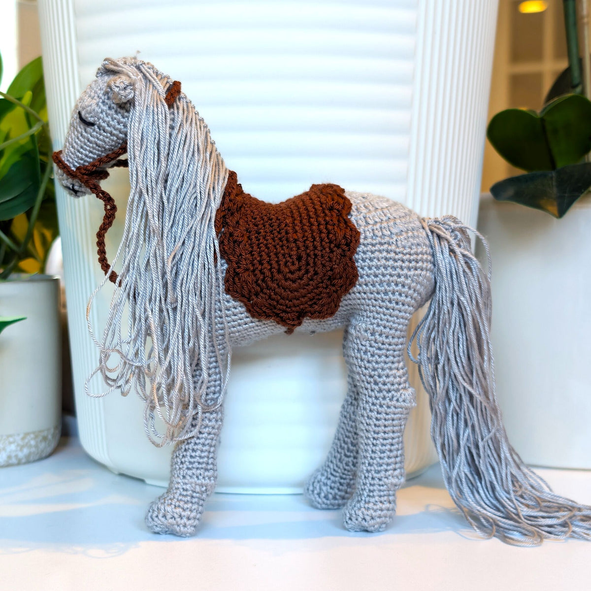 A handmade crocheted toy horse with a long gray mane and tail, brown saddle, and dark brown bridle, standing in front of a white ribbed vase with potted plants in the background.