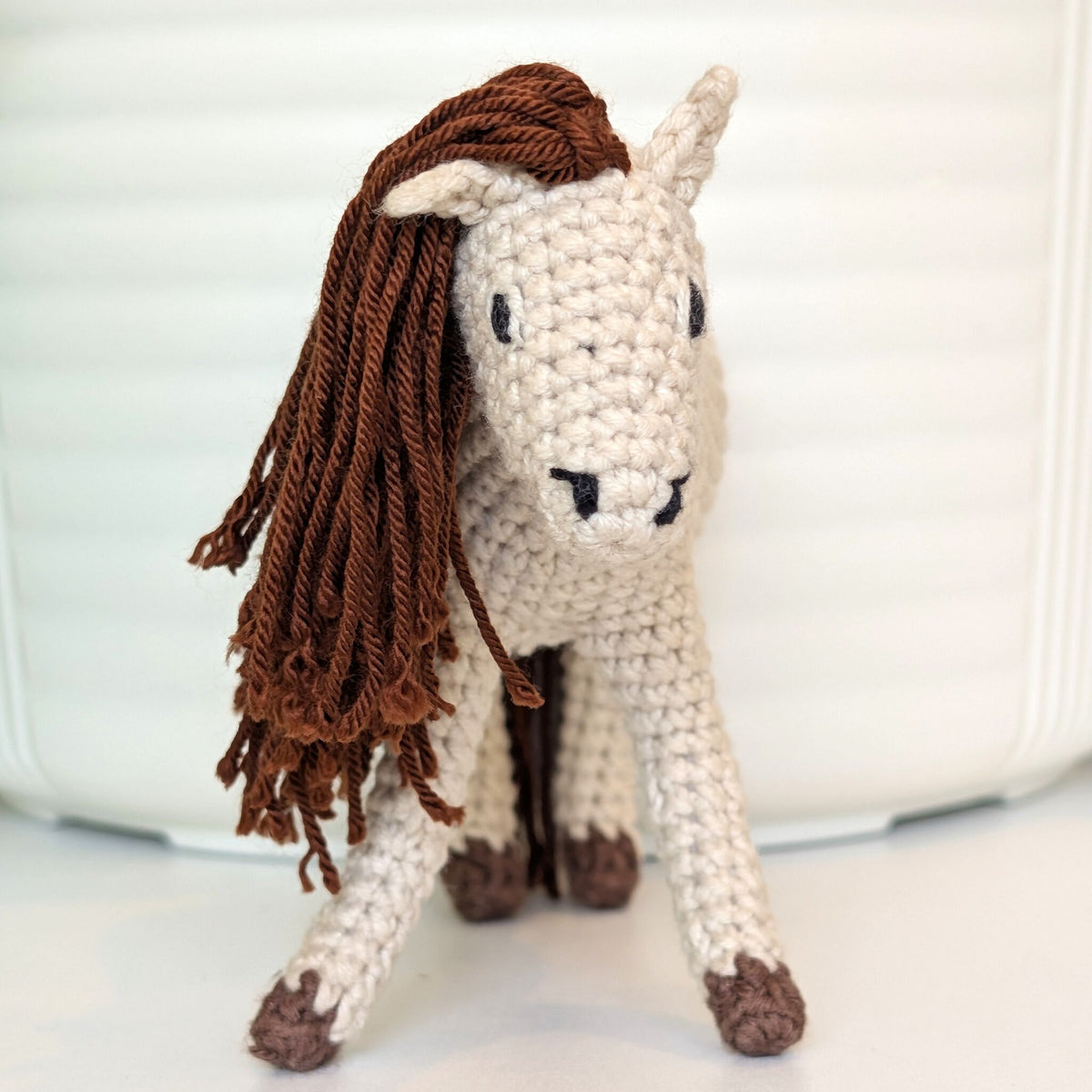 A crocheted plush horse toy with a cream-colored body and a long brown mane, standing against a white background.