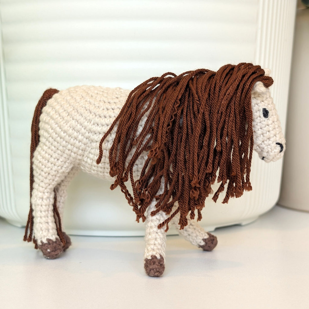 A crocheted horse figurine with a cream-colored body and a long, shaggy brown mane, standing on a white surface in front of a white ribbed container.