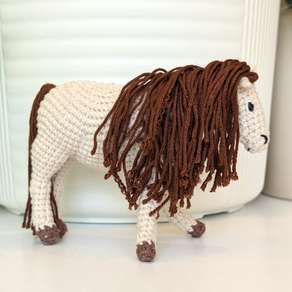 A crocheted horse figurine with a cream-colored body and a long, shaggy brown mane, standing on a white surface in front of a white ribbed container.