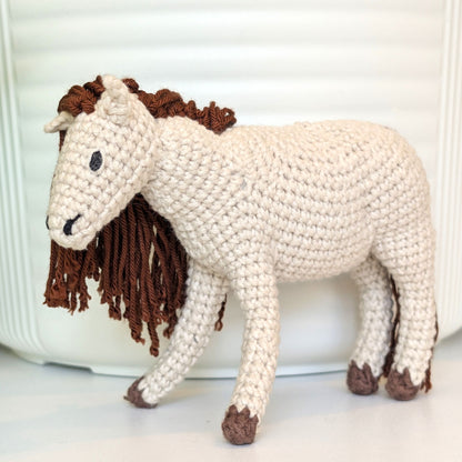 A crocheted horse figurine in off-white with a dark brown mane and tail, standing on a white surface in front of a white ribbed background.