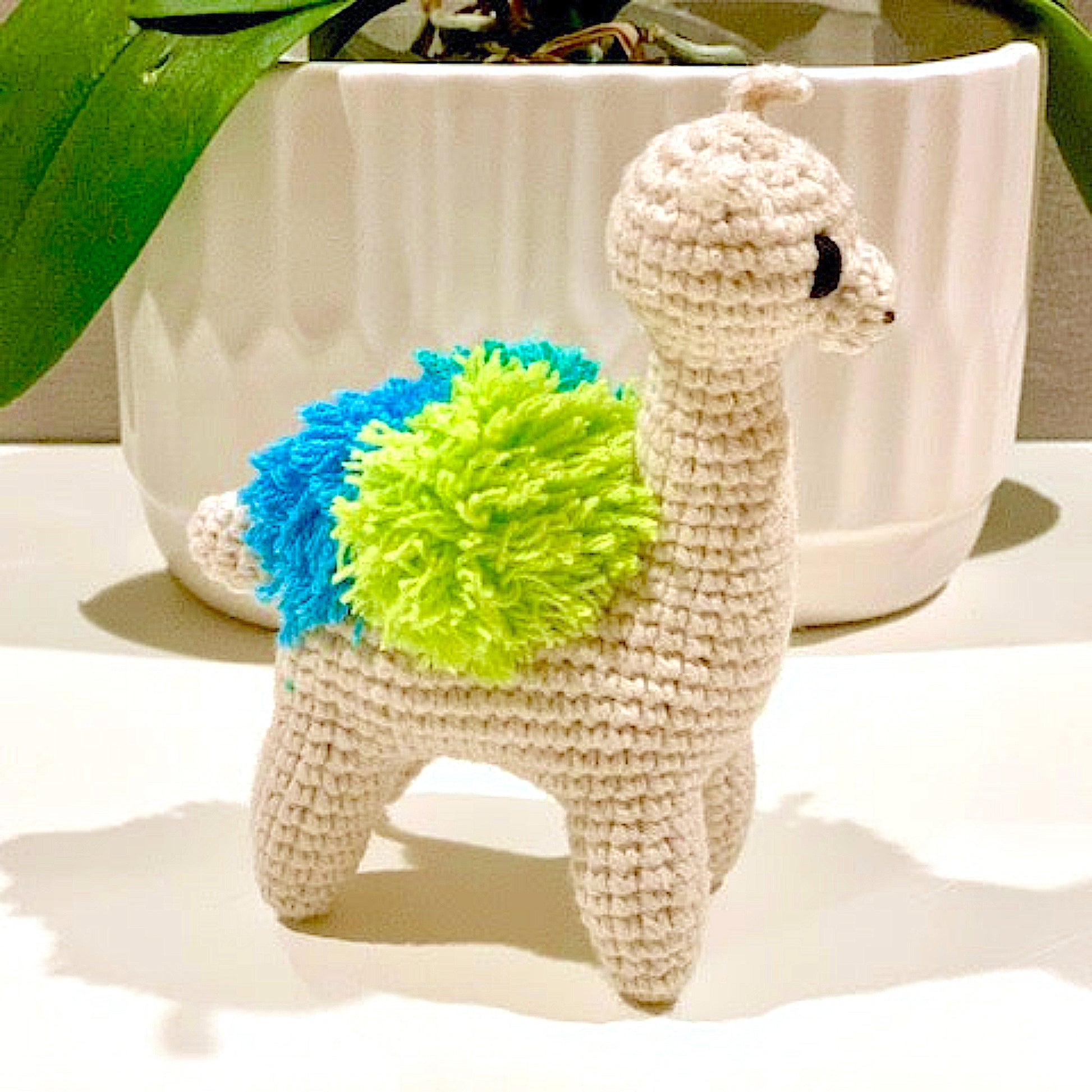 Crocheted llama toy with a blue and green accents on a white surface.