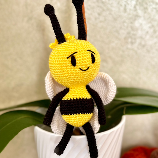 A crochet stuffed toy resembling a bumblebee with a happy facial expression, black and yellow stripes, and details for eyes and wings, sitting in a white pot with green leaves in the background.