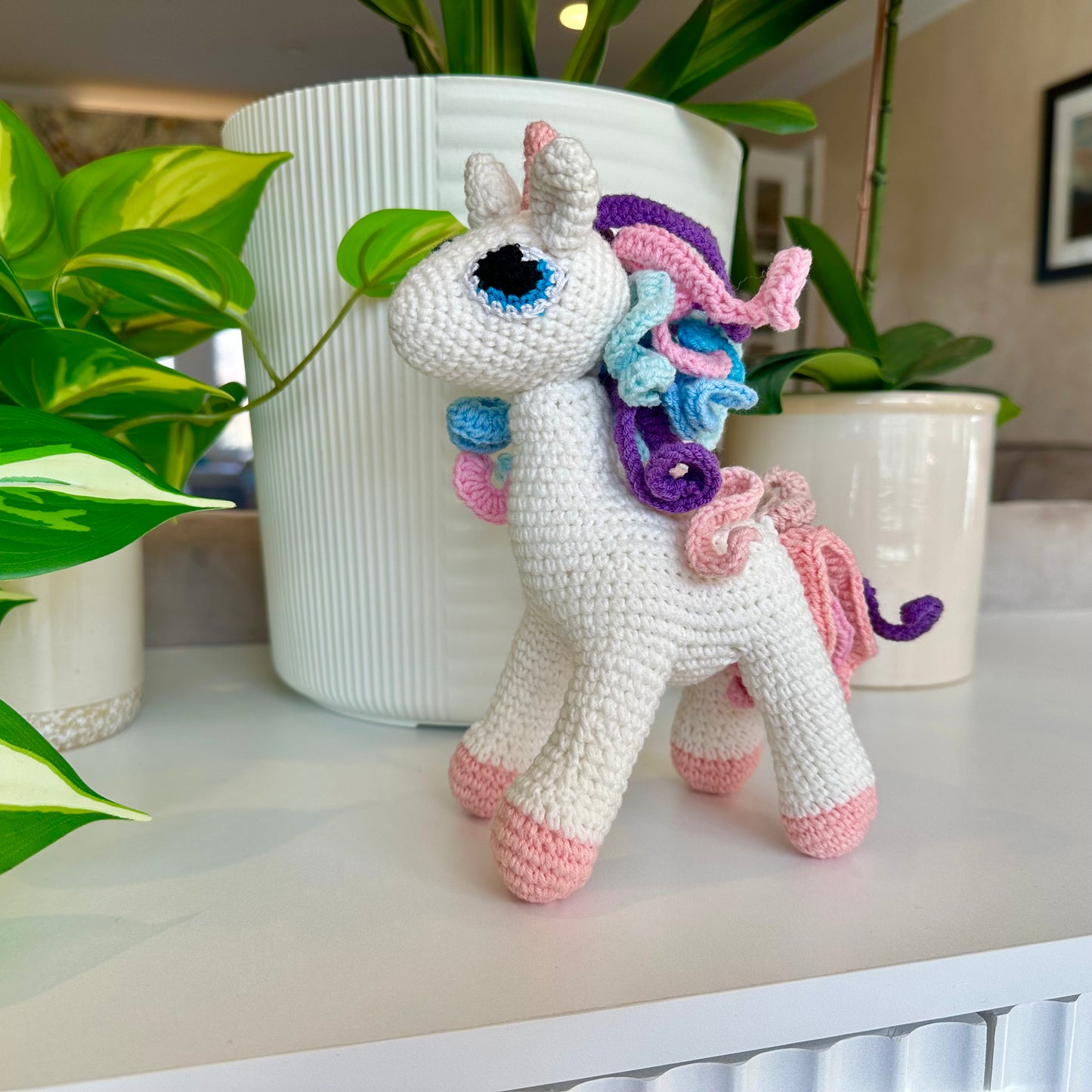 Crocheted unicorn toy with colorful mane on a surface with plants in the background
