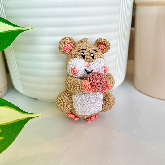 Crocheted beige hamster charm attached to a string, holding a dusty pink ball