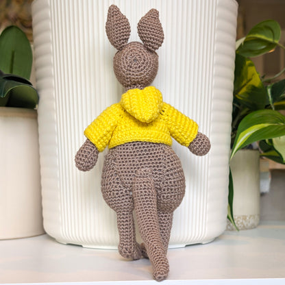 A crocheted kangaroo toy wearing a bright yellow hoodie, positioned in front of a white ribbed ceramic pot with green plants in the background.