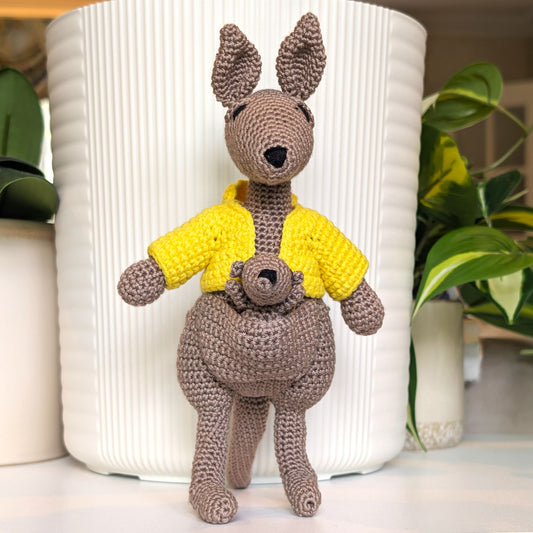 A crocheted kangaroo toy wearing a bright yellow shirt, standing in front of a white ribbed planter with green plants in the background.