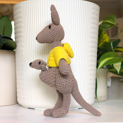 A crocheted kangaroo toy with a joey inside pouch, wearing a bright yellow shirt, posed in front of a white ribbed cylindrical planter with green plants in the background.
