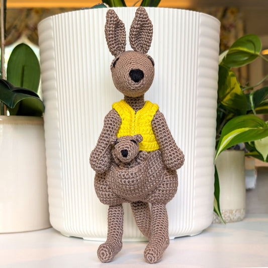 A crocheted kangaroo toy holding a small crocheted bear, wearing a bright yellow vest, posed in front of a large white ribbed planter with green plants in the background.