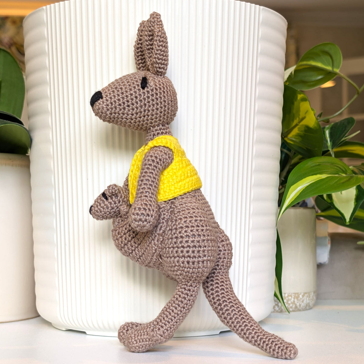 A crocheted kangaroo toy with a matching pouch holding a small kangaroo, wearing a bright yellow vest, posed in front of a white ribbed cylindrical planter with green plants in the background.