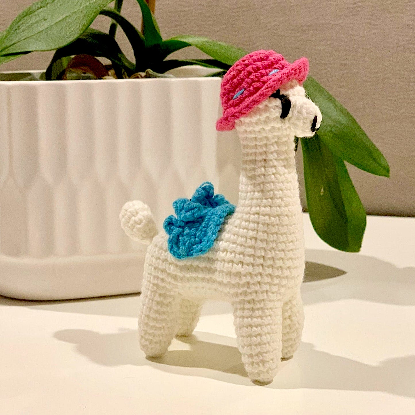 Crocheted plush llama toy with a pink hat and azure blue saddle.