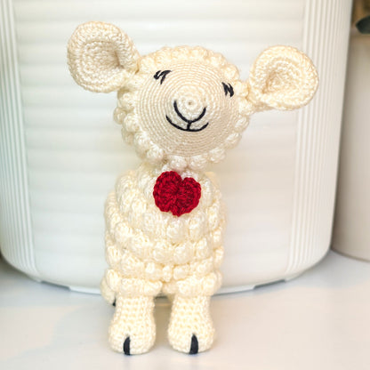 A small crocheted lamb toy with cream-colored yarn, black embroidered facial features, and a red bow tie on its chest, standing in front of a white ribbed container.