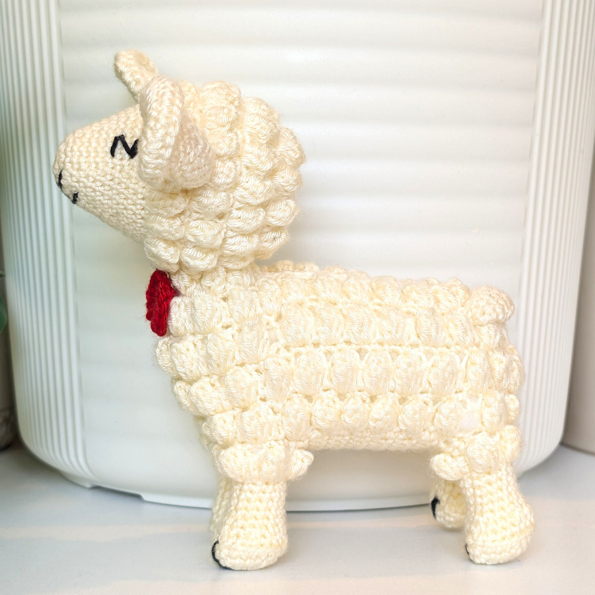 A cream-colored crocheted lamb figurine with a small red bow tie, standing in front of a white ribbed cylindrical container.