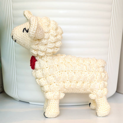A cream-colored crocheted lamb figurine with a small red bow tie, standing in front of a white ribbed cylindrical container.