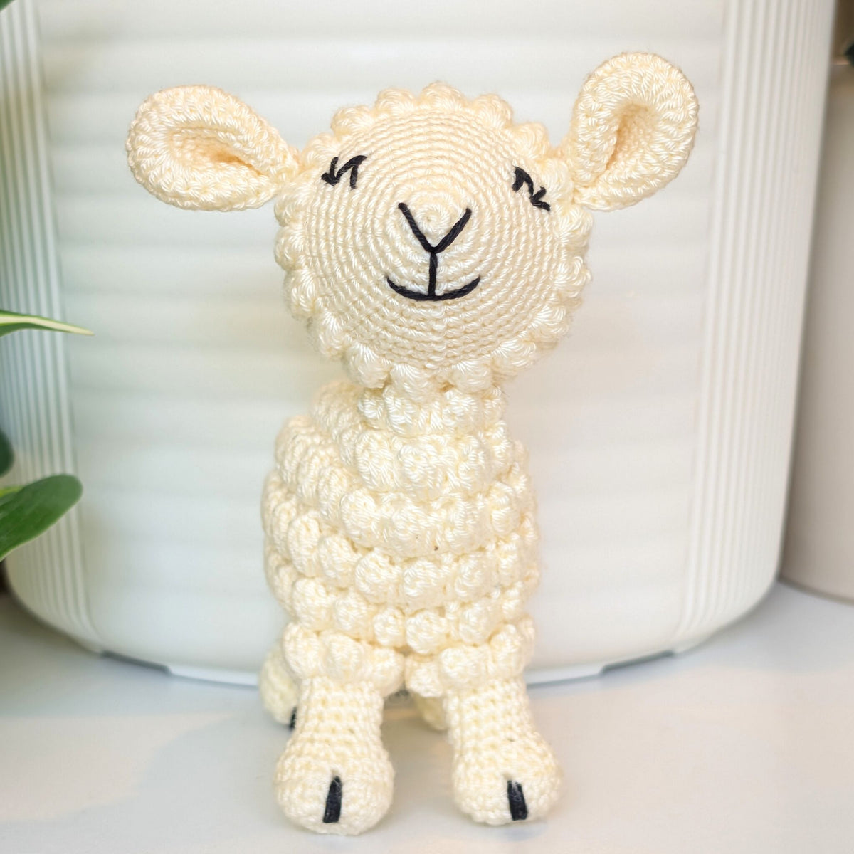 A small crocheted plush lamb toy in cream-colored yarn with black embroidered facial features, standing in front of a white ribbed ceramic pot.