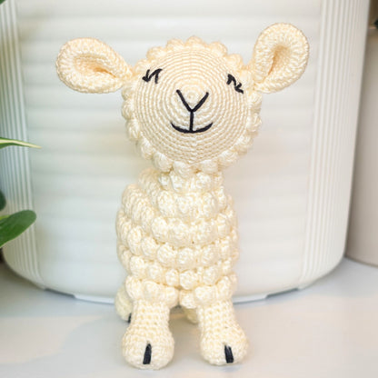 A small crocheted plush lamb toy in cream-colored yarn with black embroidered facial features, standing in front of a white ribbed ceramic pot.