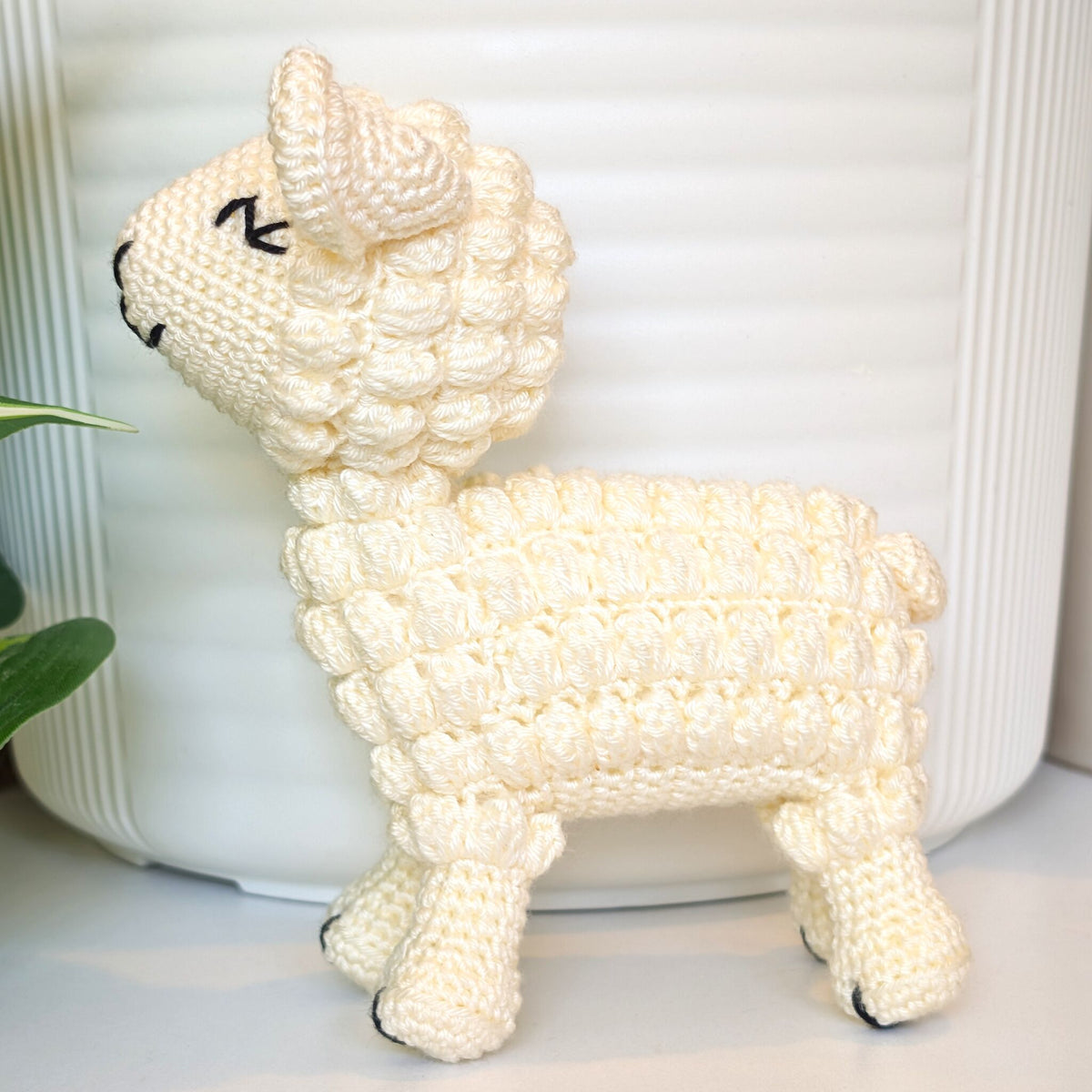 A cream-colored crocheted lamb figurine with black stitched eyes and nose, standing in front of a white ribbed ceramic pot.