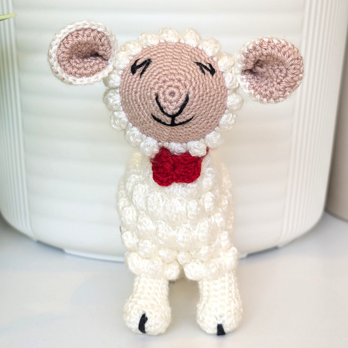 A crocheted stuffed sheep toy with fluffy white wool, a pale pink face, pink inner ears, black stitched eyes and nose, and a red bow tie, standing in front of a white ribbed container.