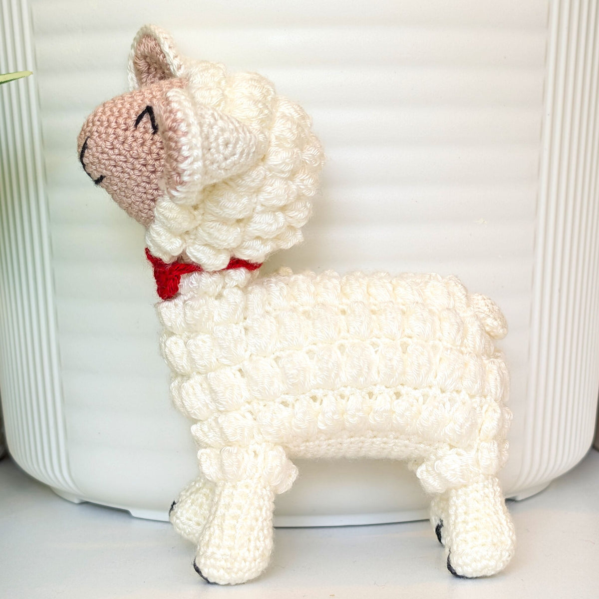 A handmade crocheted lamb toy in off-white with a pink face and a red collar, standing in front of a white ribbed background.