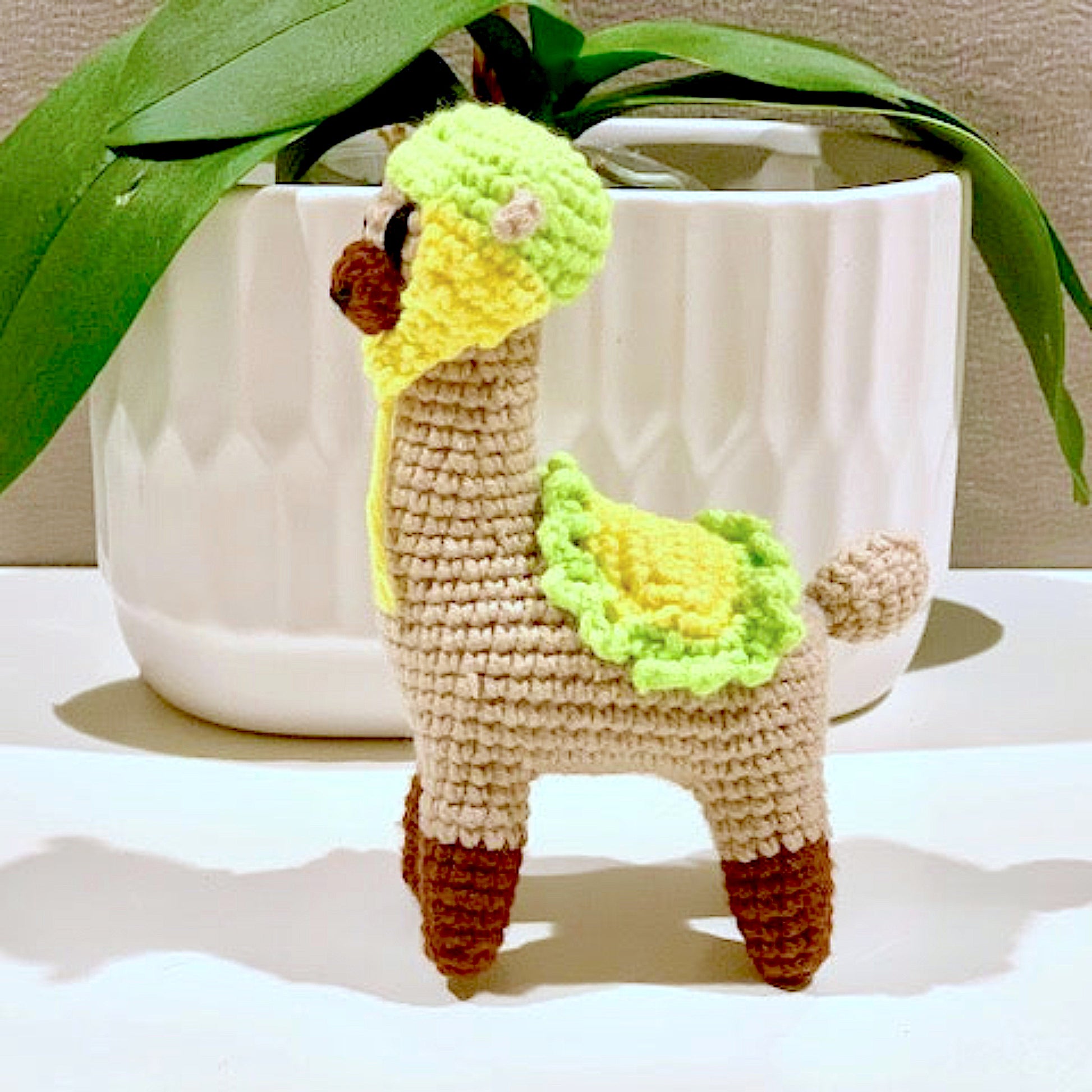 Crocheted llama toy with green accents in front of a white pot with green leaves.