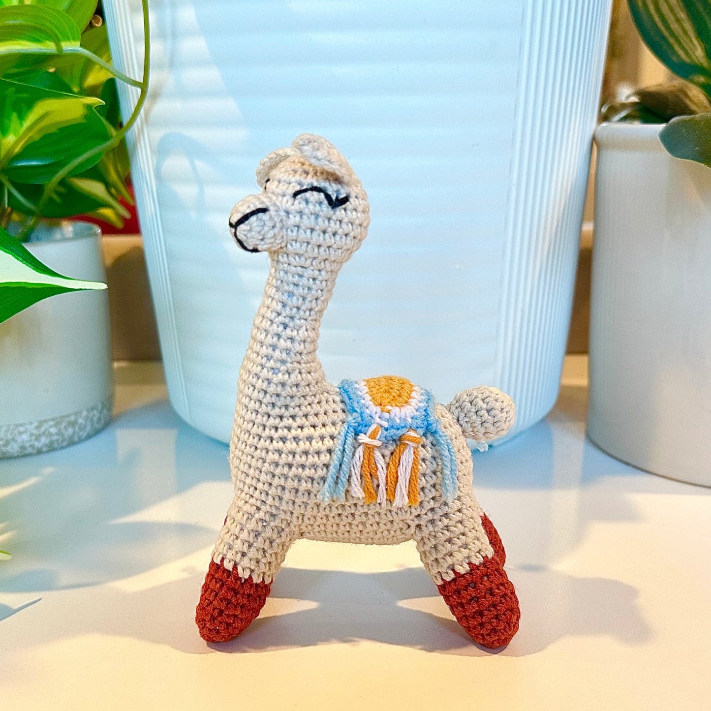 A small crocheted llama figurine with a cream body, red hooves, and a blue and yellow saddle, displayed on a white surface with potted plants in the background.